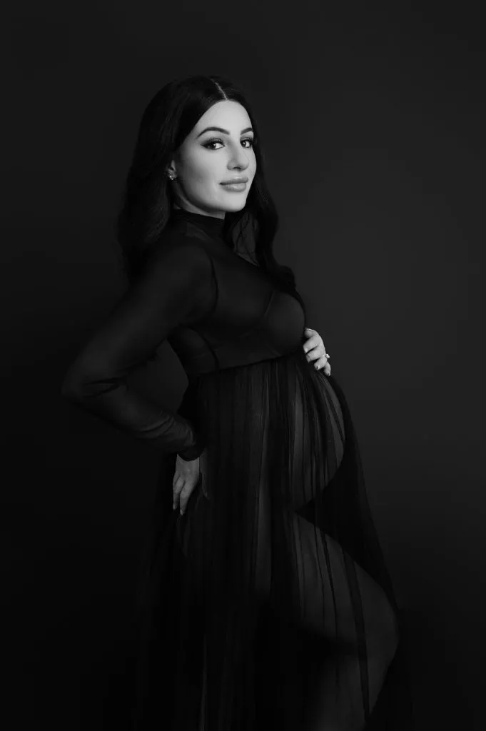 A pregnant woman poses in a sheer black dress against a dark background, one hand on her hip and the other resting on her belly, looking at the camera.