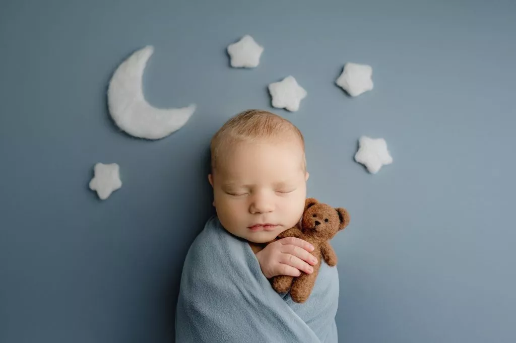 A sleeping baby wrapped in a blue blanket holds a small brown teddy bear, with felt stars and a crescent moon arranged on a blue background.