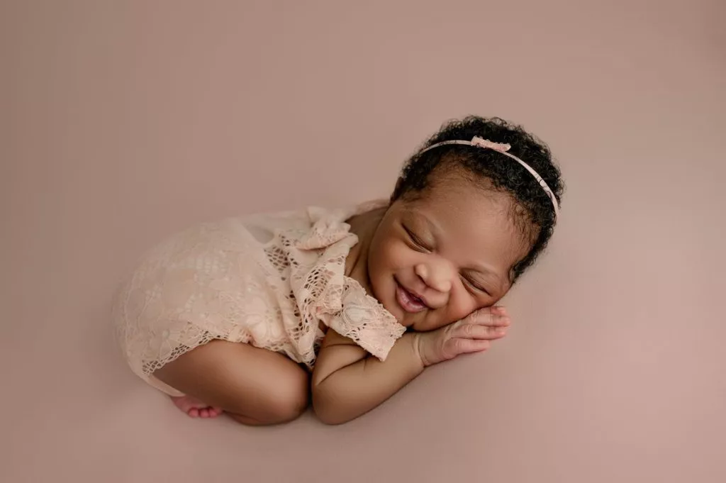 Newborn baby in a lace outfit and headband lying on a soft pink surface, smiling with eyes closed and hands under cheek.