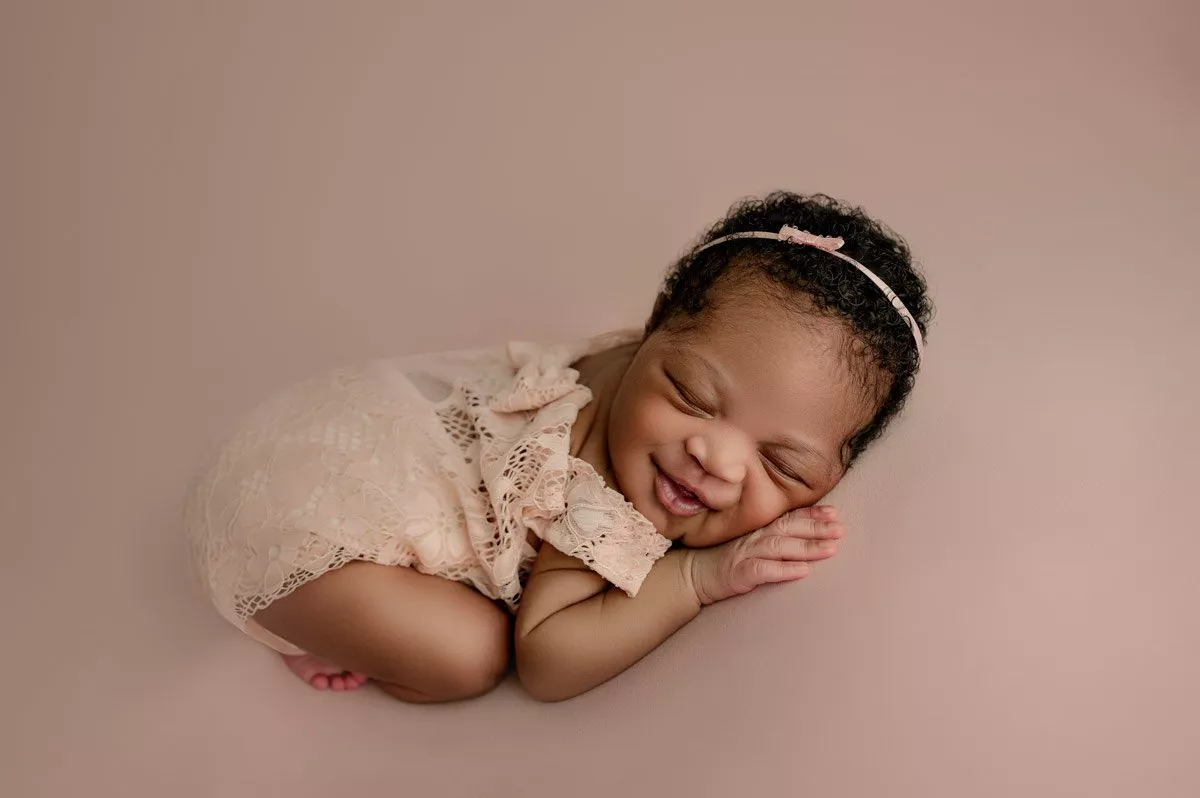 Newborn baby in a lace outfit and headband lying on a soft pink surface, smiling with eyes closed and hands under cheek.