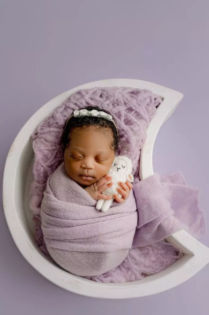Newborn baby wrapped in a lavender blanket, lying in a crescent moon crib and holding a small plush toy on a purple background—perfect for capturing those special moments in newborn photography Vancouver.