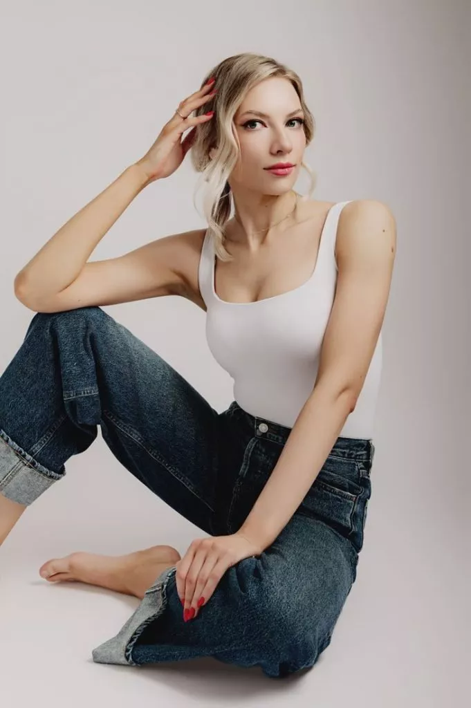 Woman with blonde hair in a white tank top and blue jeans sits on the floor against a plain background, capturing the relaxed style often seen in modern womens headshots as she looks confidently at the camera.