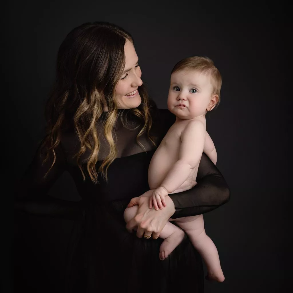 A woman in a black sheer dress holds a nude baby in her arms against a dark background. The woman is smiling at the baby.