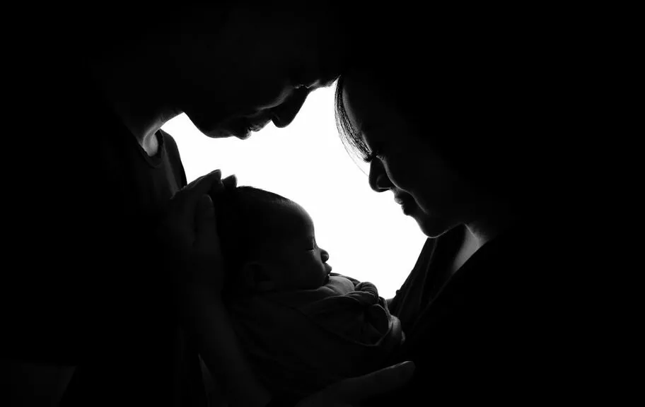 A black and white silhouette of two adults gently touching foreheads while holding an infant between them.