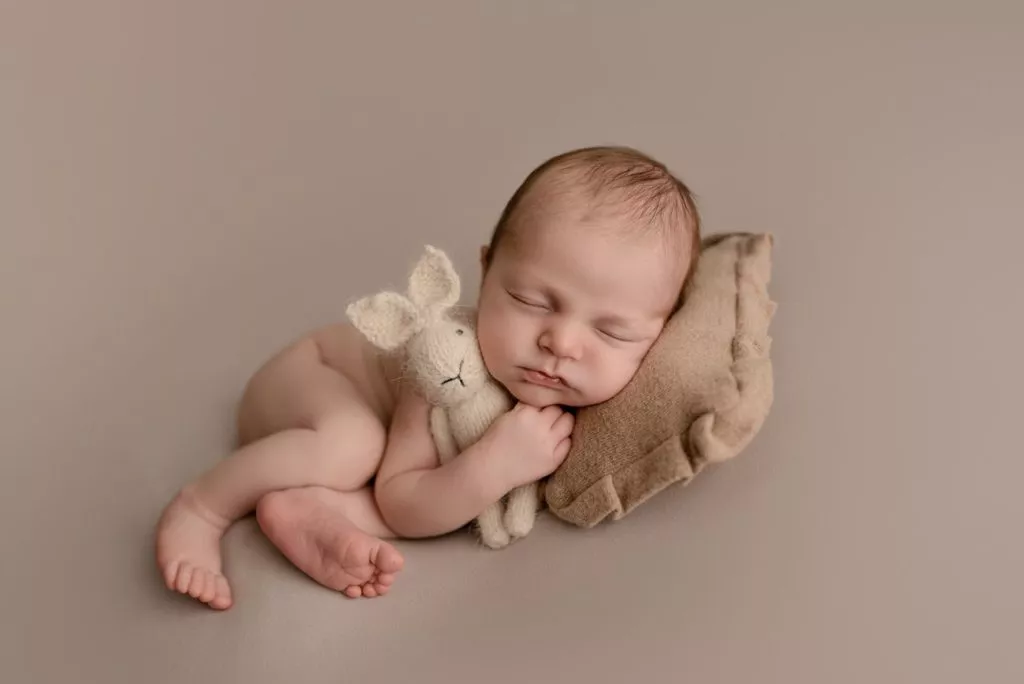 A newborn baby sleeps on a beige pillow, hugging a small knitted bunny toy, against a plain beige background in a cozy Burnaby setting.