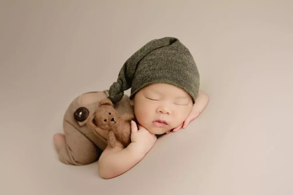 A newborn baby wearing a green hat and brown outfit sleeps on a beige surface, holding a small brown teddy bear—a heartwarming moment captured in North Vancouver.
