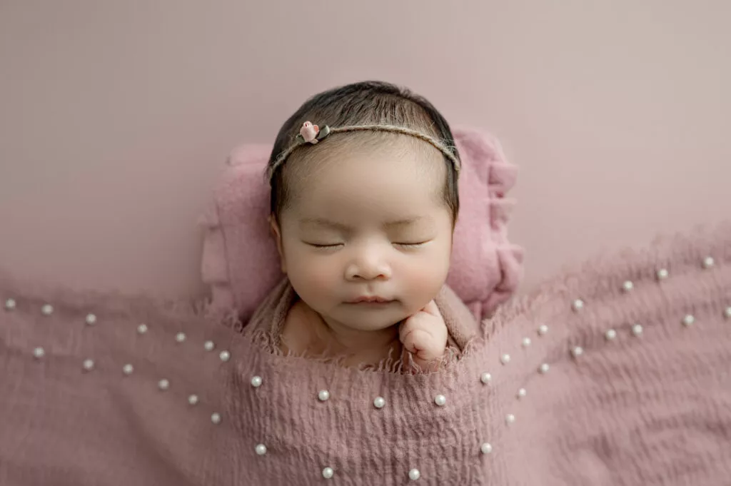 A newborn baby sleeps on a pink blanket with a matching headband and a textured, pearl-decorated covering, capturing the sweetness of Newborn Photography Vancouver.