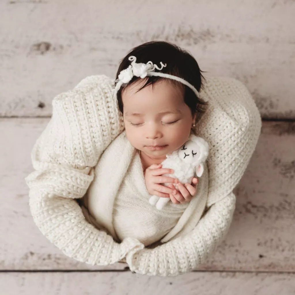 A baby wearing a white knitted outfit and headband sleeps while holding a small plush toy, lying on a light wooden surface, perfectly capturing the gentle innocence of Newborn Photography Vancouver.