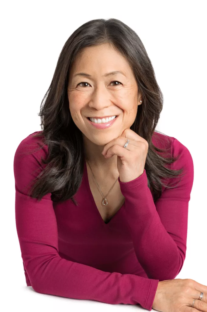 Woman with long dark hair, wearing a magenta top and silver necklace, smiling and resting her chin on her hand against a white background—perfect for professional womens headshots.