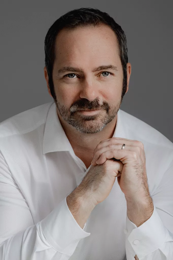 A man with short dark hair and a beard, wearing a white dress shirt, sits with his hands clasped and looks directly at the camera against a gray background—a style often seen in professional womens headshots.
