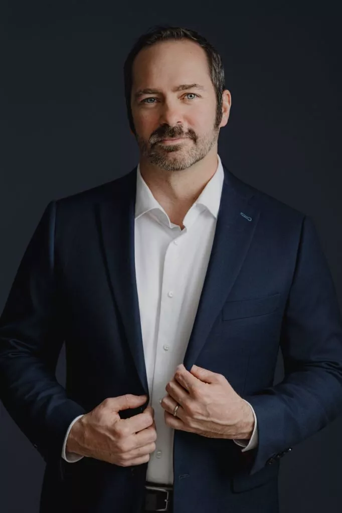 A man in a dark suit and white shirt stands against a dark background, looking at the camera with a neutral expression, hands adjusting his jacket—a professional pose often seen in womens headshots as well.