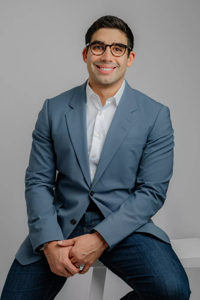 A man wearing glasses, a blue blazer, white shirt, and jeans sits on a white stool against a plain gray background, smiling at the camera.