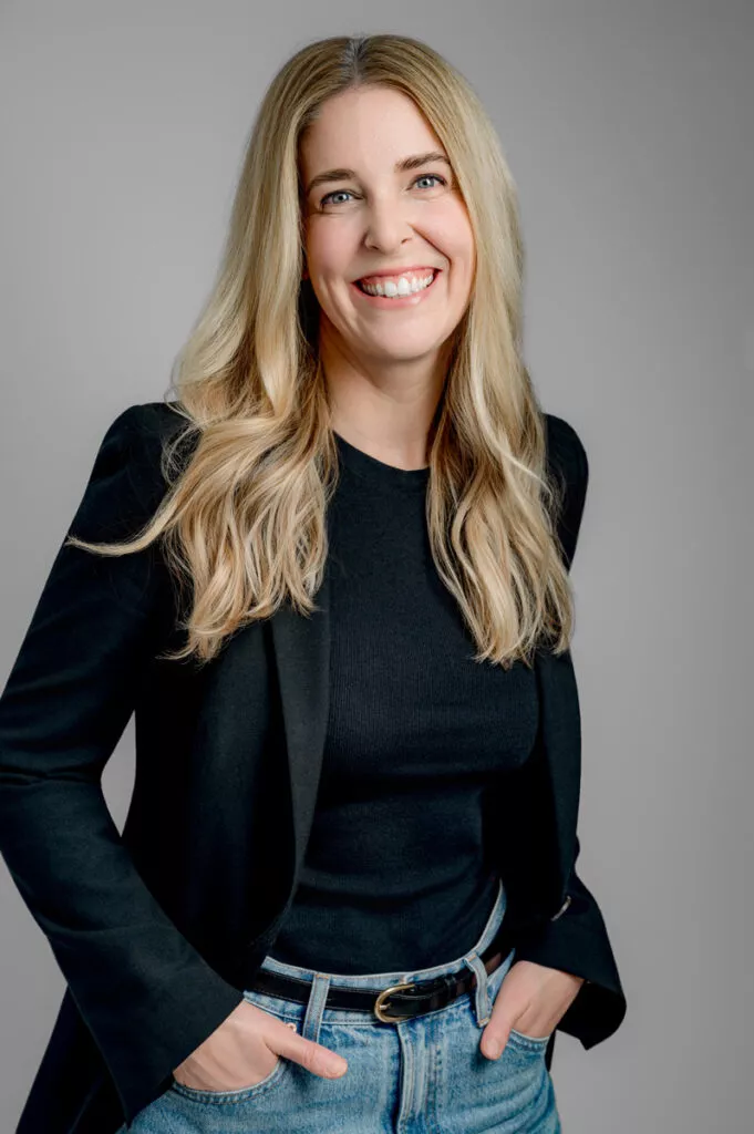 A woman with long blonde hair wearing a black top, black blazer, and blue jeans stands smiling with her hands in her pockets against a plain gray background.