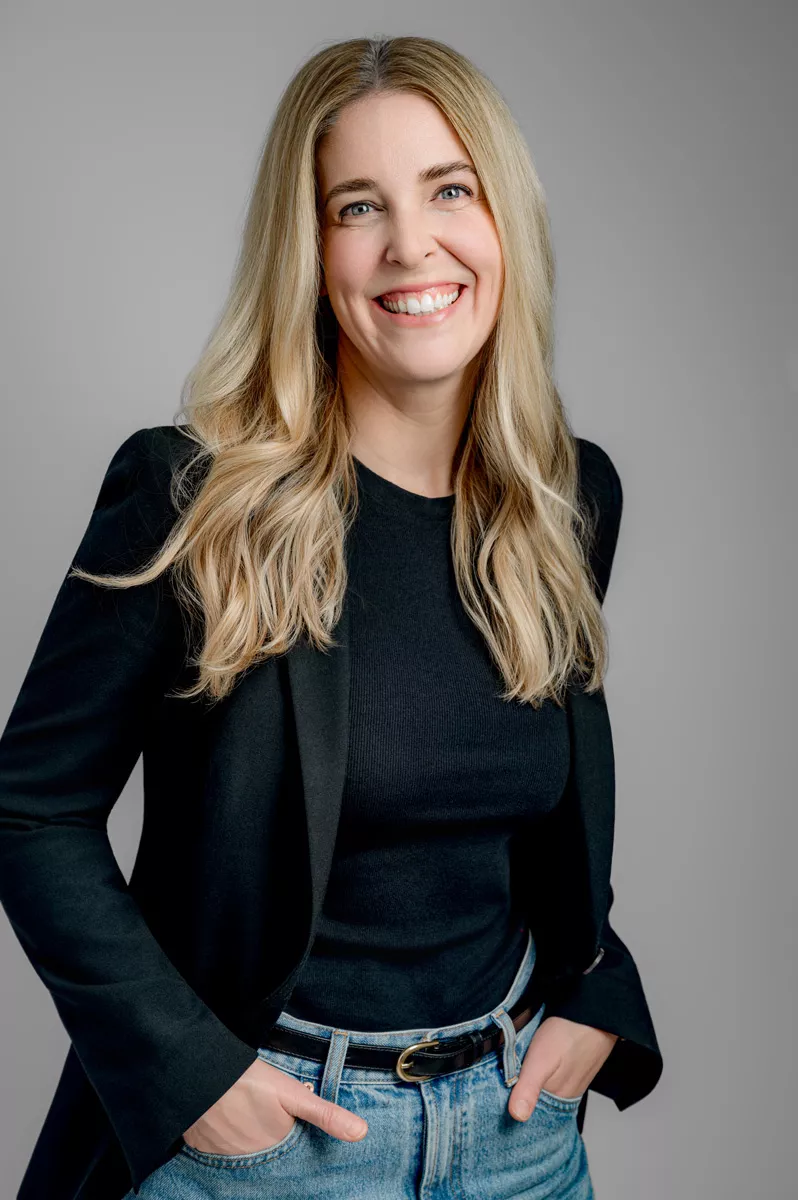 A woman with long blonde hair wearing a black top, black blazer, and blue jeans stands smiling with her hands in her pockets against a plain gray background.