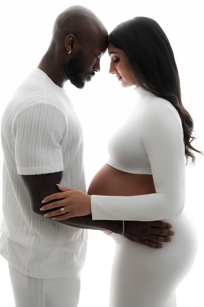 A man and a pregnant woman stand face to face, gently touching foreheads and holding her belly, both dressed in white against a white background—a serene moment captured by T Haller Photography, specialists in newborn photography Vancouver.