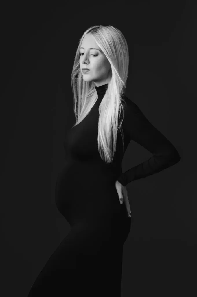 A pregnant woman with long blonde hair poses in profile against a dark background, wearing a fitted black dress and resting one hand on her back—a timeless maternity portrait by T Haller Photography, specialists in newborn photography Vancouver.