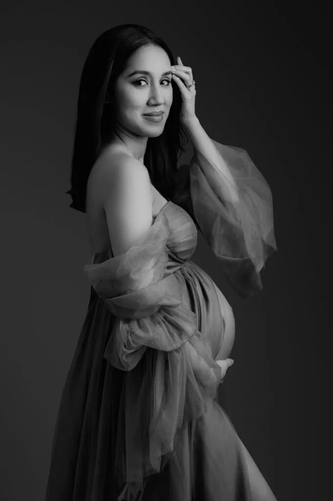 Black and white portrait by T Haller Photography of a pregnant woman in a sheer, flowing dress, posing gracefully with one hand on her belly—a timeless moment captured by this newborn photography Vancouver artist.