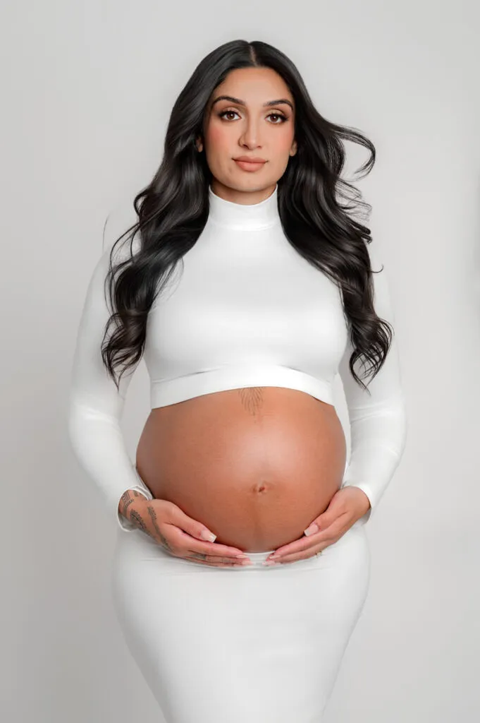 A pregnant woman with long dark hair, wearing a fitted white outfit, stands against a plain light background with her hands resting on her bare belly—captured by T Haller Photography, specialists in newborn photography Vancouver.