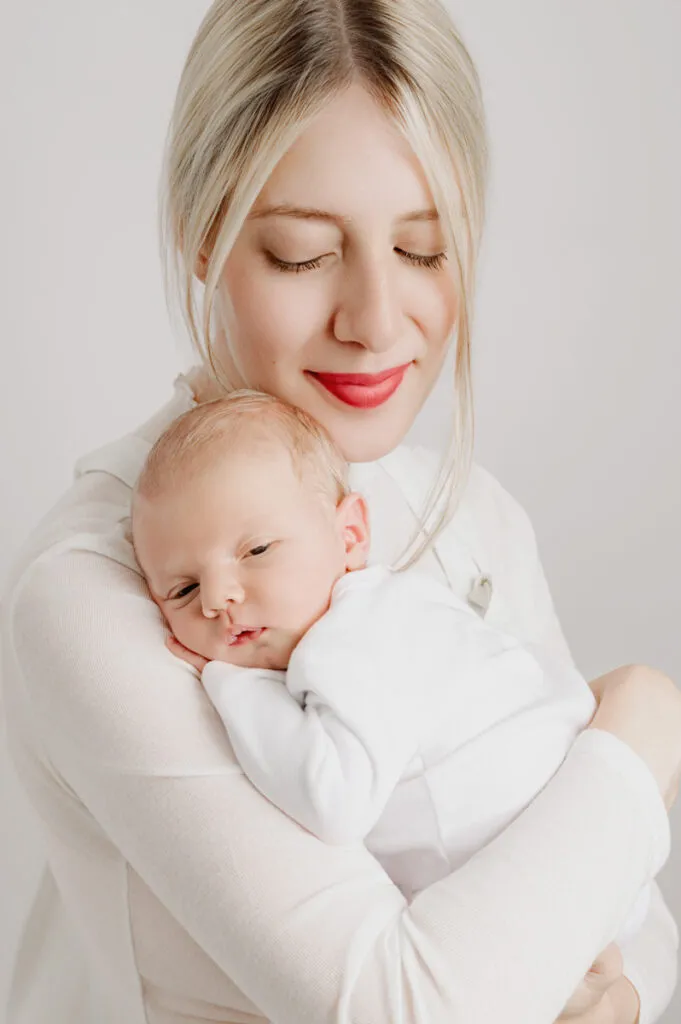 A woman with blonde hair holds a sleeping baby close to her chest. Both are dressed in white, set against a light and neutral background—an image that beautifully captures the tender moment by a skilled newborn photographer.
