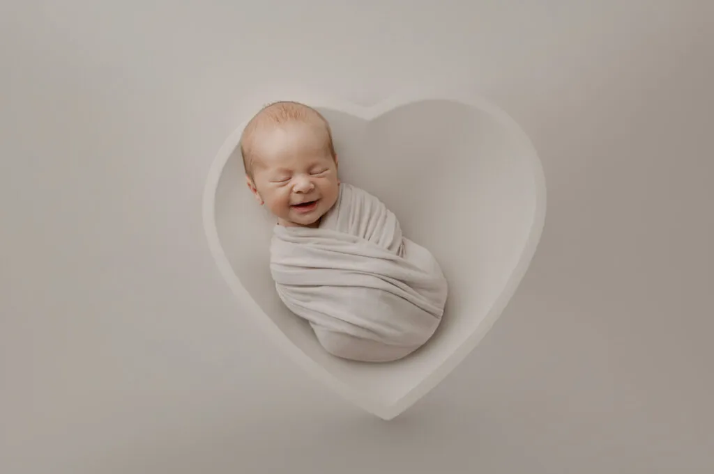 A newborn baby wrapped in a white blanket is lying in a white, heart-shaped bowl with a neutral background, perfectly styled for a newborn photographer’s portfolio.
