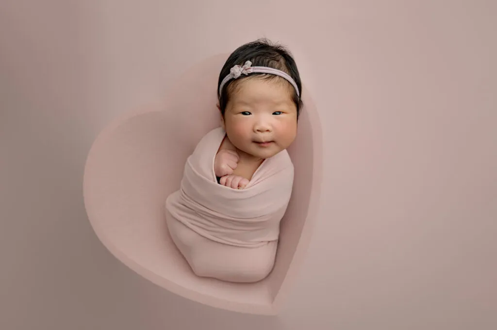 A newborn photographer captures a baby wrapped in a pink blanket, lying in a heart-shaped cushion, with a pink headband, against a pale pink background.