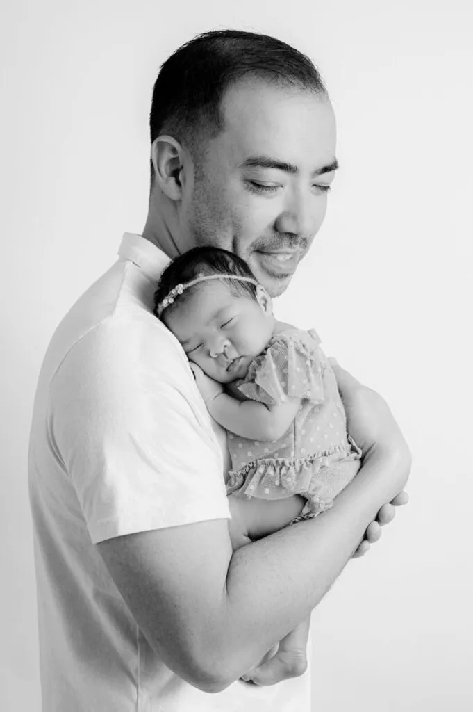 A man holding a sleeping baby girl close to his chest in a gentle embrace, both facing slightly to the side, captured by a skilled newborn photographer against a plain background.