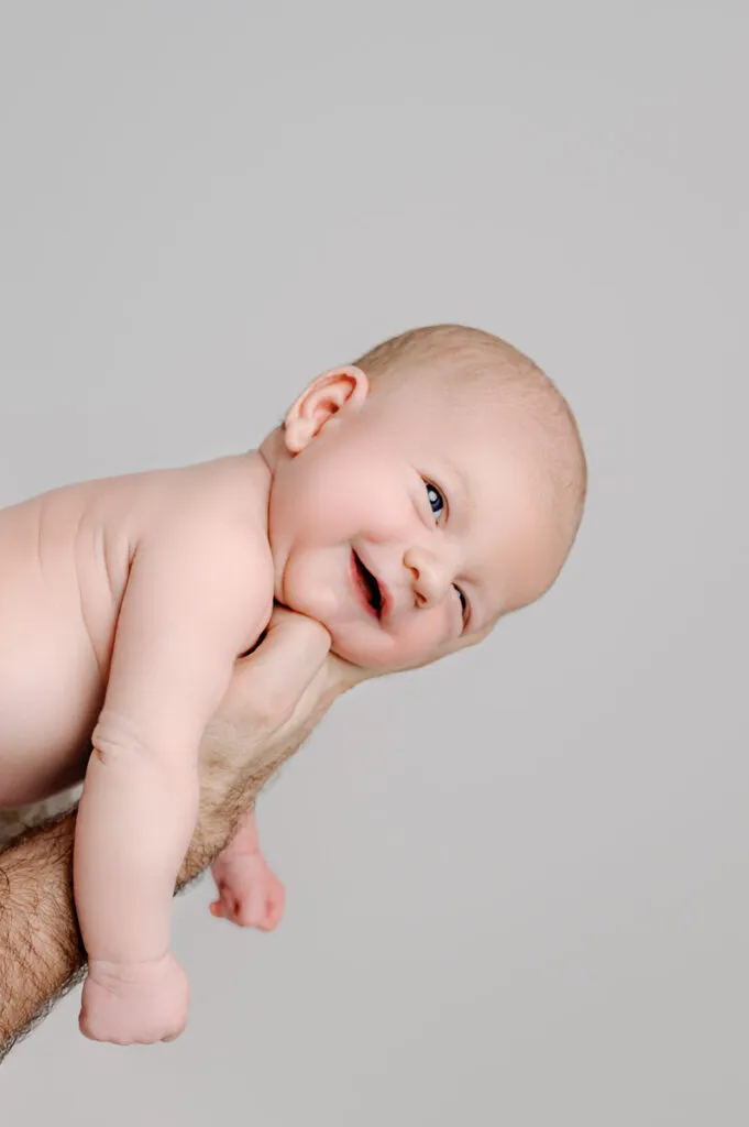 An adult hand holds a smiling baby, gently posed on their stomach against a plain light gray background—a perfect moment for a newborn photographer to capture.