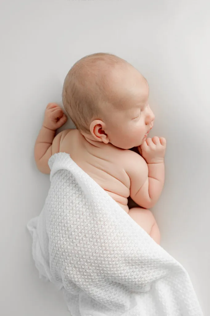 A baby lies on its side on a white surface, partially covered with a white textured blanket, eyes closed and hands near its face—a serene moment often captured by a skilled newborn photographer.