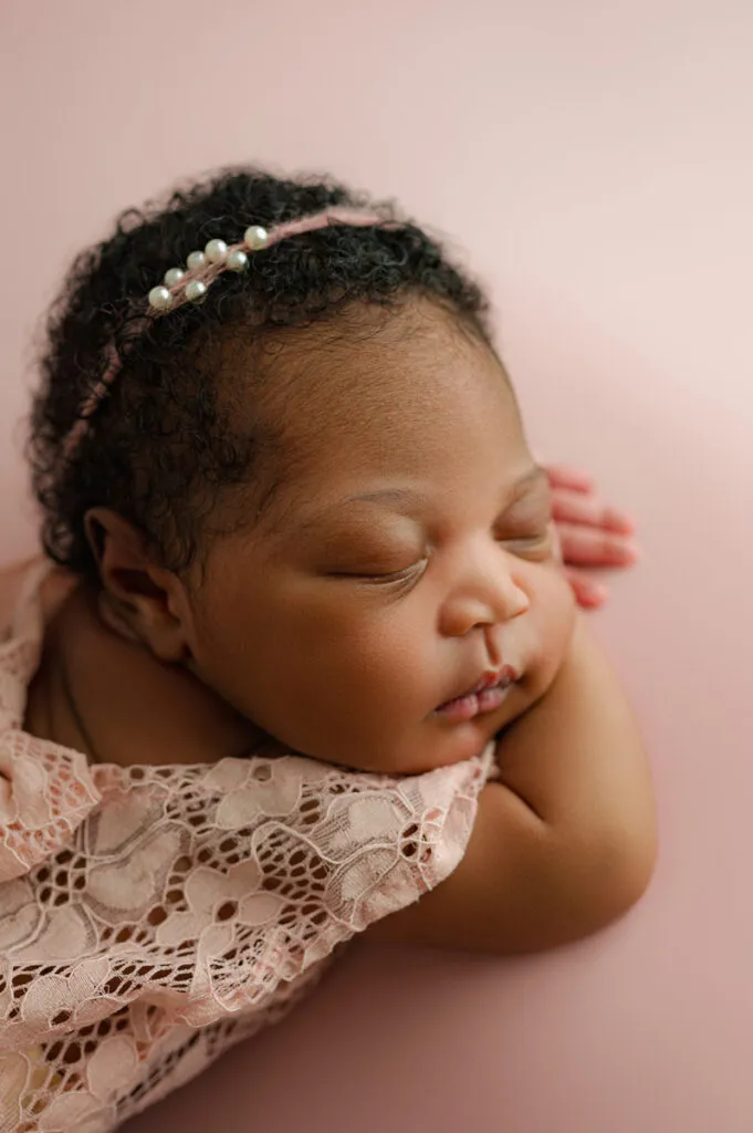 A newborn photographer captures a baby in a lace outfit and pearl headband, peacefully sleeping on a soft pink surface with tiny hands under the cheek.