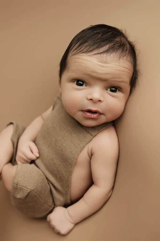 A baby with dark hair and brown eyes, captured by a newborn photographer, sits in a brown sleeveless outfit on a matching background, looking up at the camera.