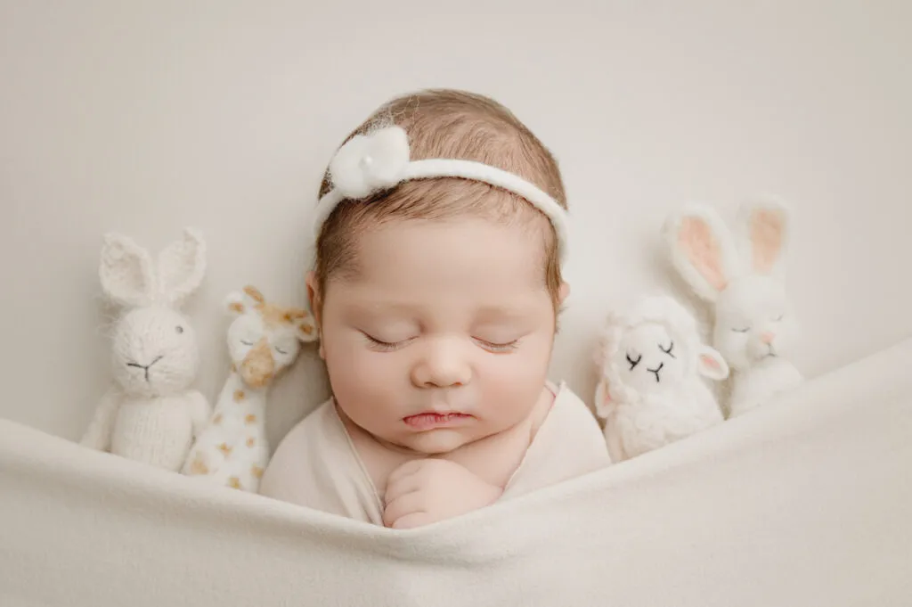 A sleeping baby with a white headband is tucked under a blanket, surrounded by four stuffed animal toys on a light background—an adorable scene captured perfectly by a skilled newborn photographer.