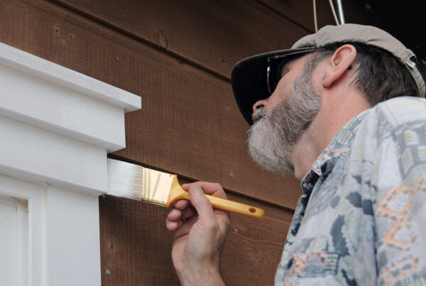 Exterior painting in progress as a handyman uses a brush to apply white paint to exterior window trim on a wooden house.