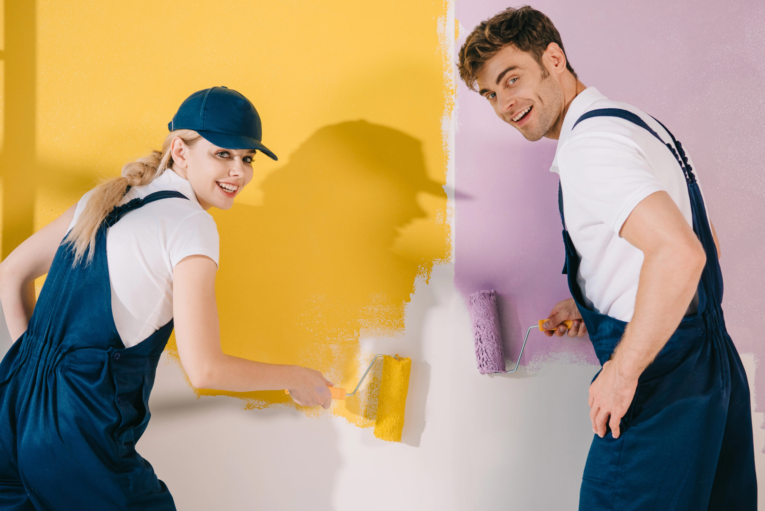 Interior painting services in action with two painters applying yellow and lavender paint to an indoor wall using rollers.