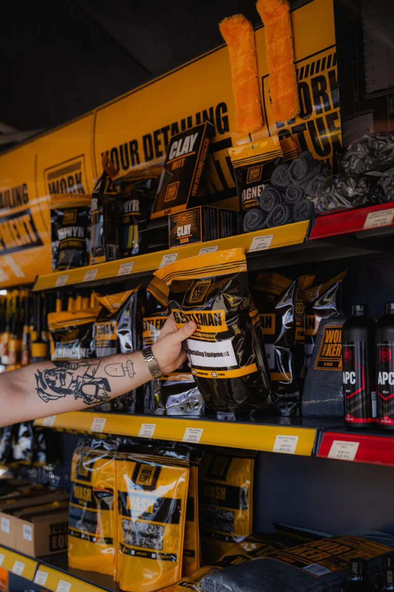 A tattooed arm grabs a GENTLEMAN package from a West Hall shelf with yellow and black auto detailing supplies nearby.