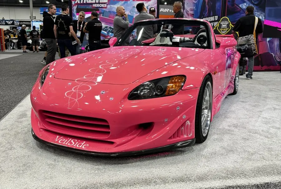 A bright pink VeilSide Honda S2000 at SEMA Show 2025, crowds and colorful booths with automotive banners behind.