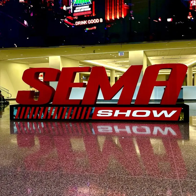 Large red 3D SEMA SHOW sign on a shiny marble floor indoors, with digital screens and city images in the background.