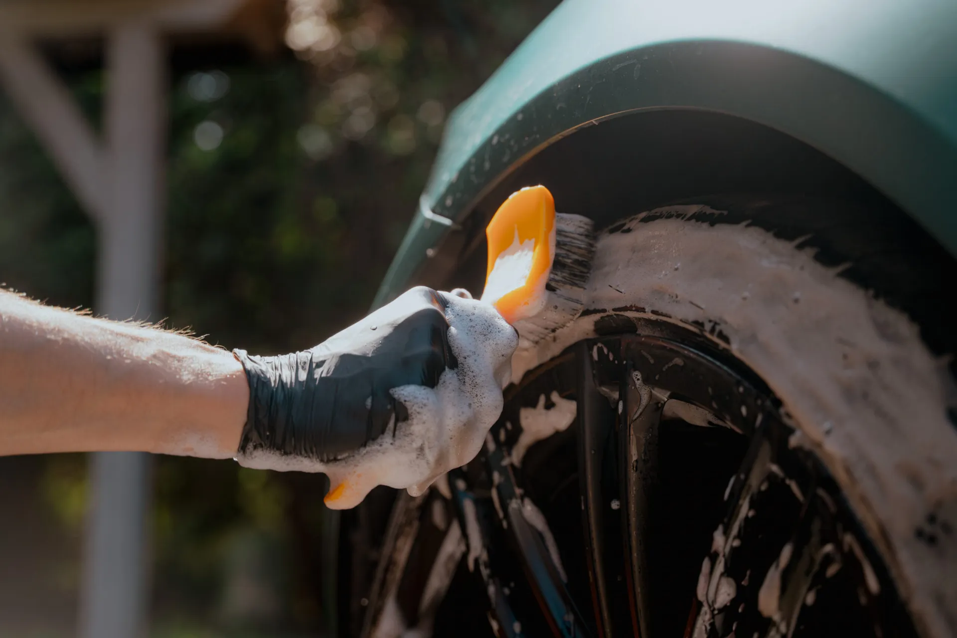 A black-gloved hand uses a Work Stuff Willy Willy orange-handled detailing tool to scrub soap onto a car tire and wheel.