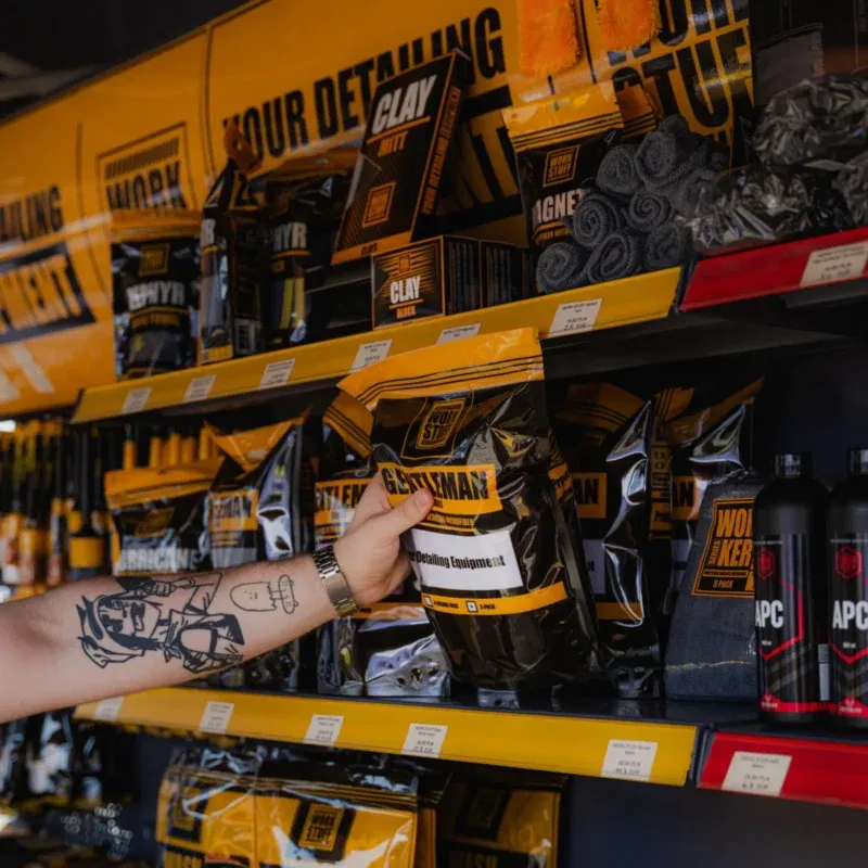 A tattooed arm reaches for a GENTLEMAN package on a shelf filled with car detailing products in yellow and black packaging.
