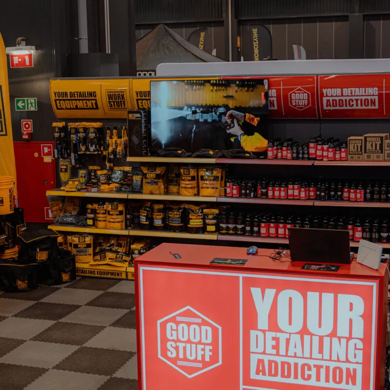 Trade show booth with car detailing kits, yellow buckets, and bottles on a large red Good Stuff counter; TV and banners visible.