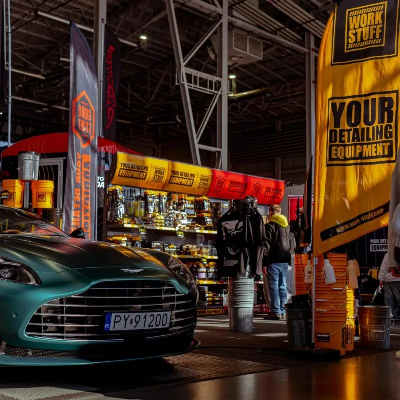 Front of a green car with license plate at AutoCosmetica 2026, yellow orange banners, Work Stuff shelves, people browsing.