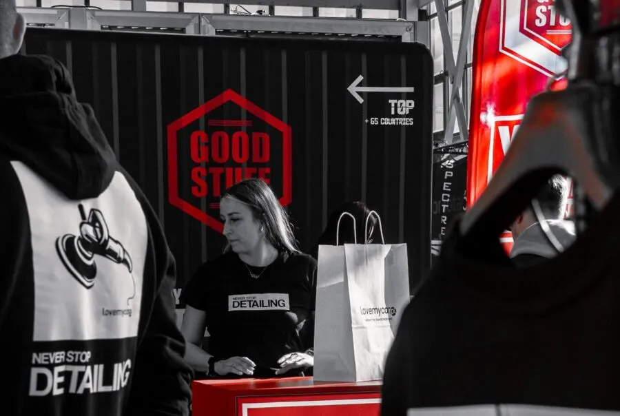 A woman in a Never Stop Detailing shirt stands behind a red counter with shopping bags; Work Stuff Good Stuff container visible.