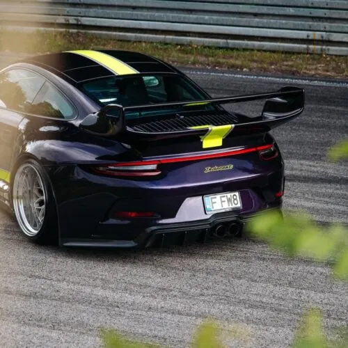 Coche deportivo morado oscuro con rayas de carreras amarillas y alerón trasero aparcado junto a un quitamiedos metálico, matrícula F F M 8.