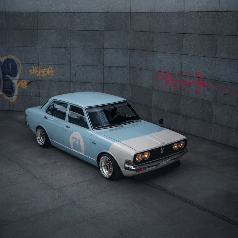 A vintage light blue sedan with number 71 on the door is parked on gray tiles, near tall concrete walls with red graffiti.