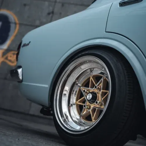 Rueda trasera de coche antiguo azul claro con llanta multirradio dorada y neumático de perfil bajo junto a un muro de hormigón con grafitis.