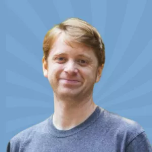 A person with short light brown hair, wearing a gray t-shirt, is smiling in front of a blue, sunburst-patterned background.