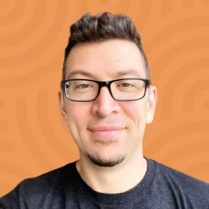 A person with short dark hair and glasses is smiling at the camera. They are wearing a dark T-shirt. The background is an orange pattern with circular lines.