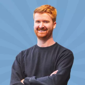 A person with red hair and a beard stands against a blue background with radiating lines, wearing a black sweater and smiling with arms crossed.