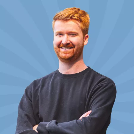 A person with red hair and a beard stands against a blue background with radiating lines, wearing a black sweater and smiling with arms crossed.