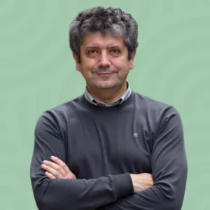 A person with curly hair and a slight smile stands with arms crossed, wearing a gray sweater over a collared shirt. The background is light green with a subtle circular pattern.