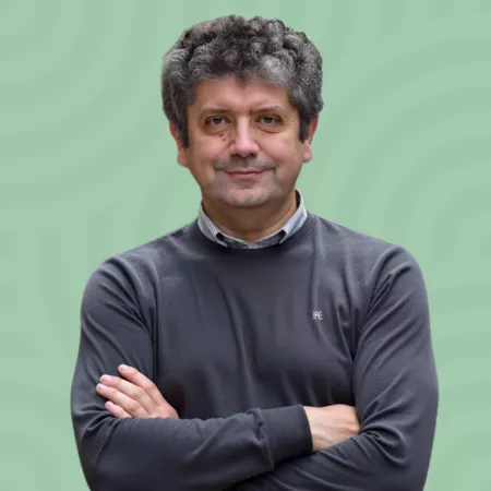 A person with curly hair and a slight smile stands with arms crossed, wearing a gray sweater over a collared shirt. The background is light green with a subtle circular pattern.